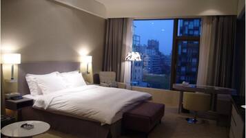 Habitación de hotel con una cama grande, dos lámparas de noche, una mesita pequeña, una silla y vistas a la ciudad desde la ventana.