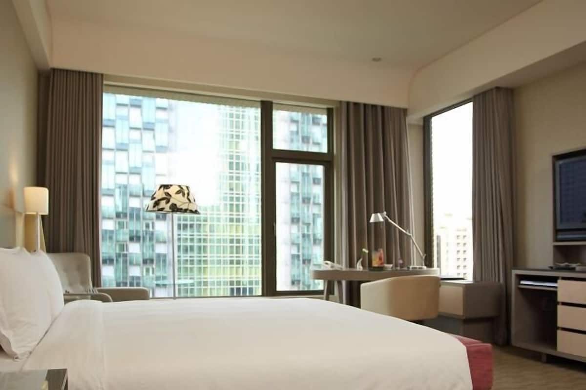 Habitación de hotel con cama, escritorio, silla, lámpara y una ventana grande con vista a la ciudad.