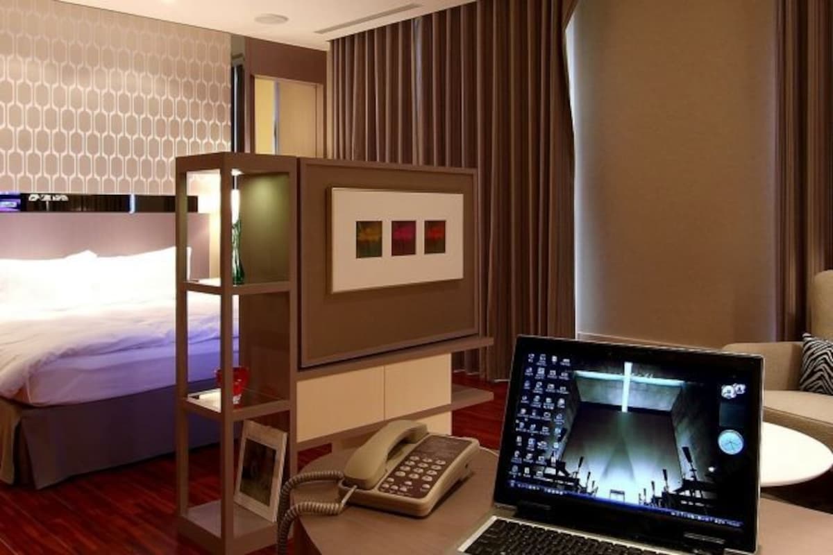 A hotel room with a bed, a desk with a laptop, a telephone, and a wall-mounted TV.