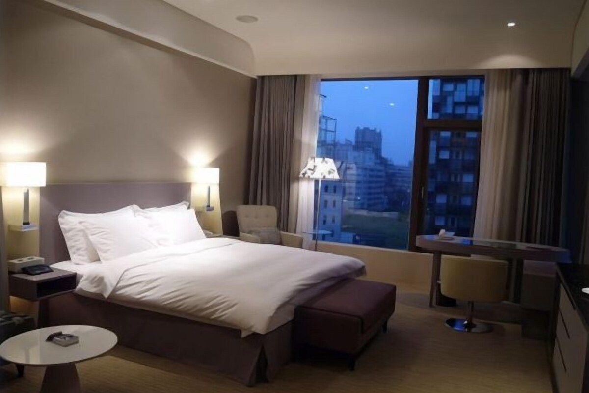 Habitación de hotel con una cama grande, mesitas de noche, una pequeña mesa redonda, una silla y vistas a la ciudad.