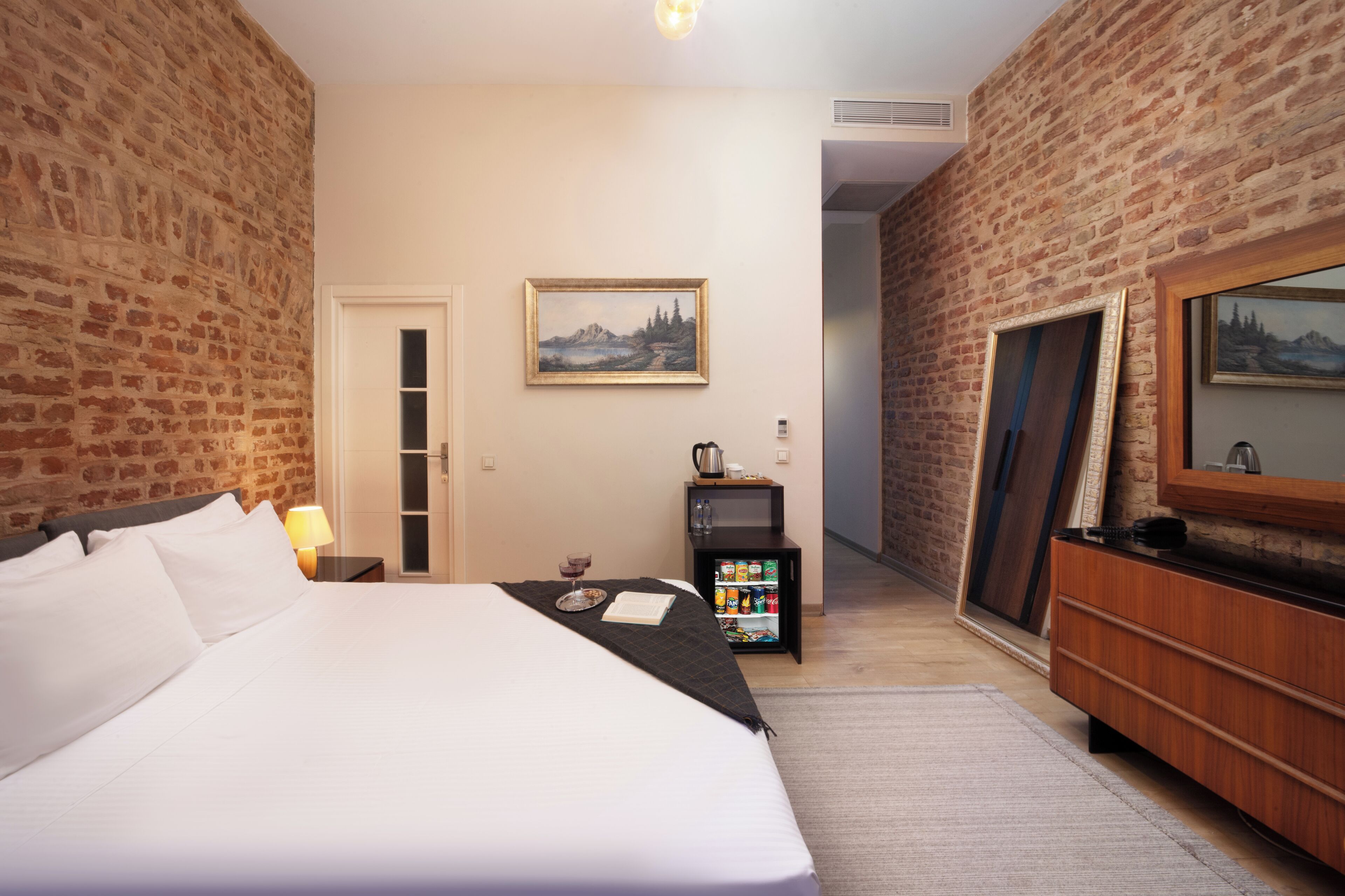 Habitació d'hotel amb un llit gran, una taula petita amb una làmpada, un quadre a la paret, un minibar i un mirall.