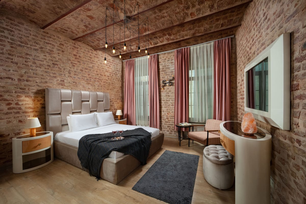 Une chambre avec un mur en briques, un lit avec une tête de lit, une table de chevet, une chaise, une table et une grande fenêtre avec des rideaux.