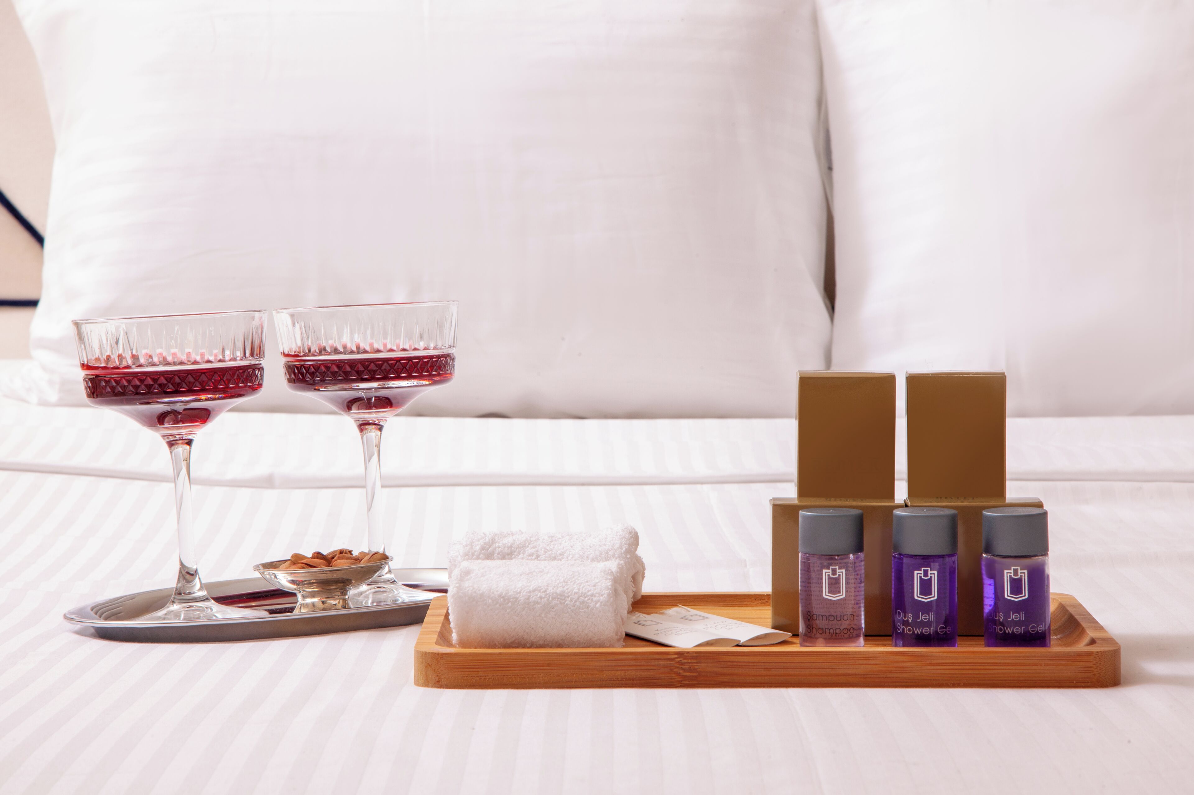 A neatly arranged tray with two glasses of red liquid, a bowl of almonds, a folded white towel, and three bottles of shampoo and shower gel on a bed with white pillows.