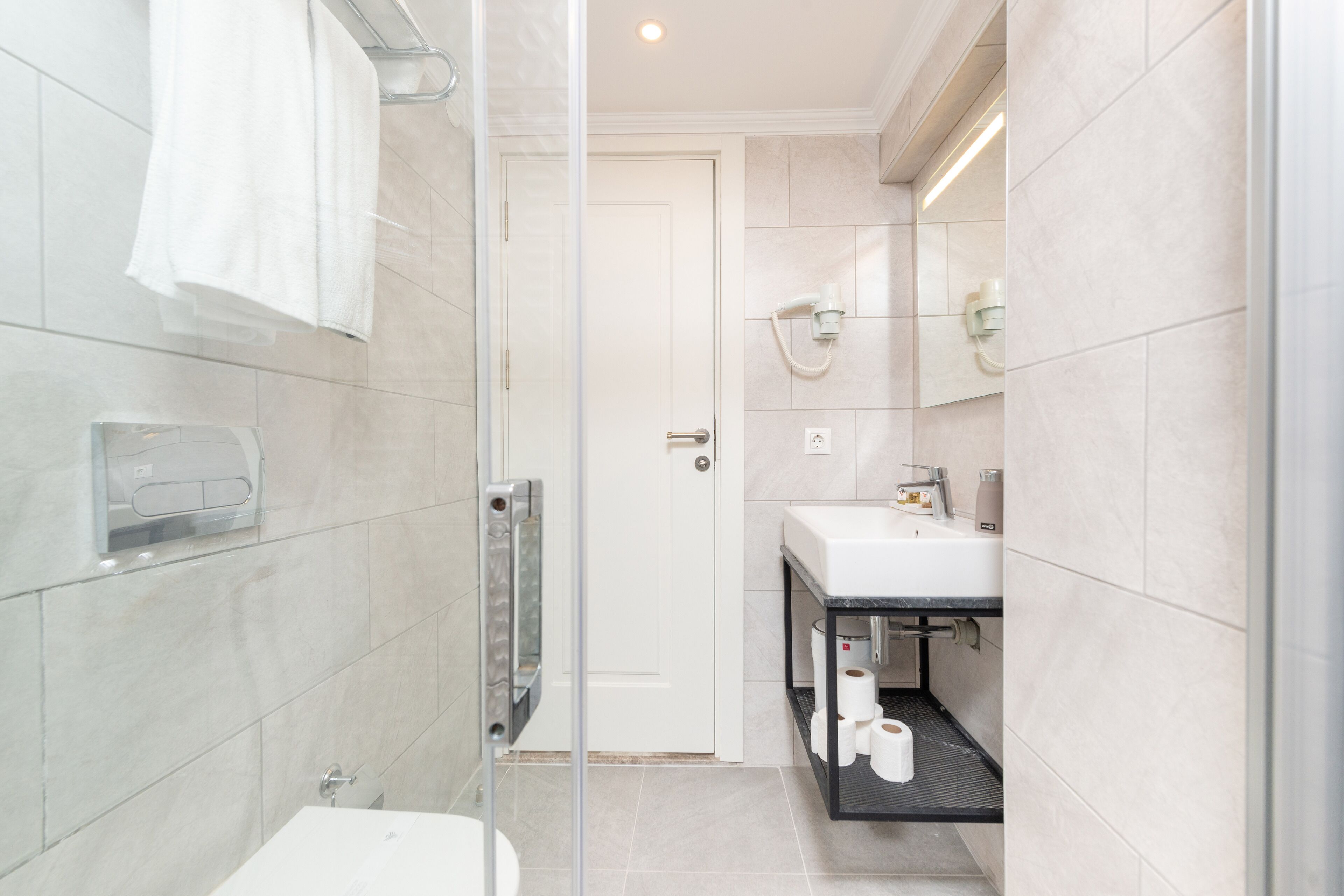 A modern bathroom with a glass shower enclosure, a toilet, a sink with a mirror, and a shelf with toilet paper.