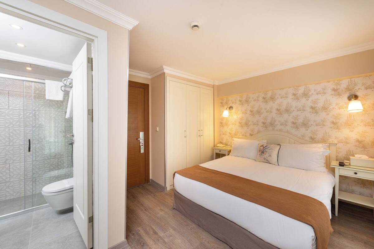 Habitación de hotel con una cama grande, una mesita de noche y un baño con cabina de ducha de vidrio, además de una puerta que da a otra habitación.