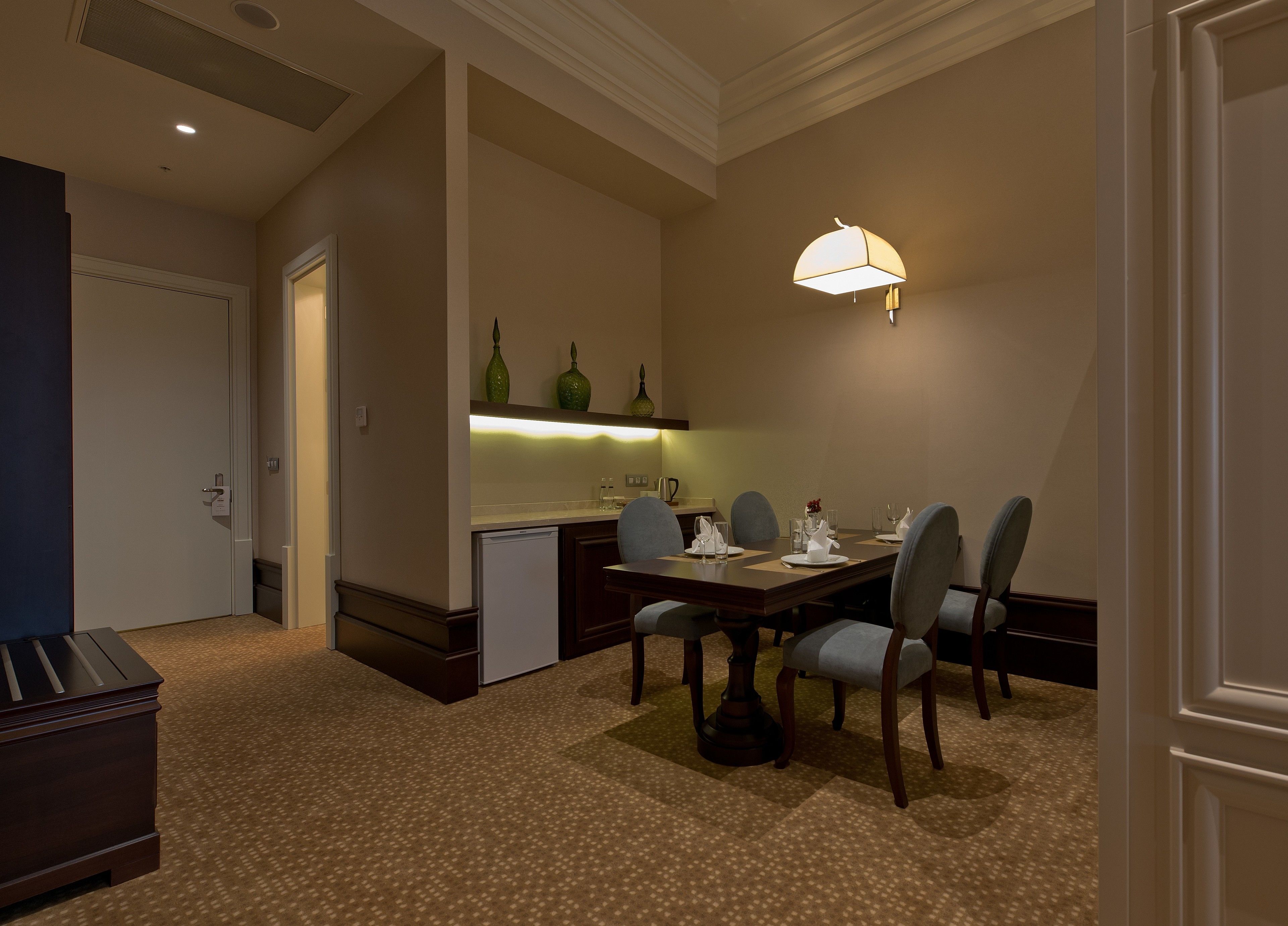 Comedor de hotel con una mesa dispuesta para cuatro, un refrigerador, un microondas y una lámpara fijada a la pared.