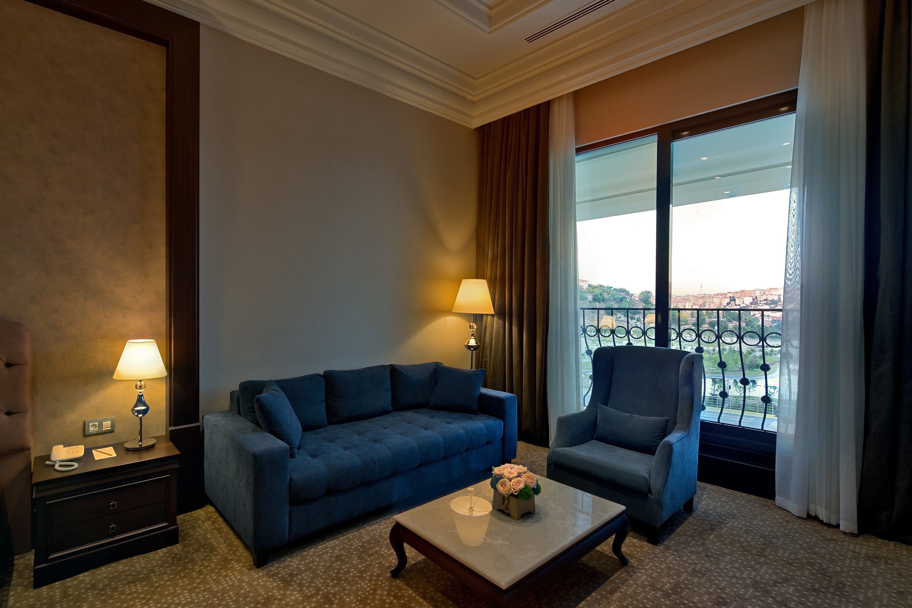 A hotel room with a blue sofa, a matching armchair, a small table with a vase of flowers, a nightstand with a lamp, and a large window with a view of a cityscape.