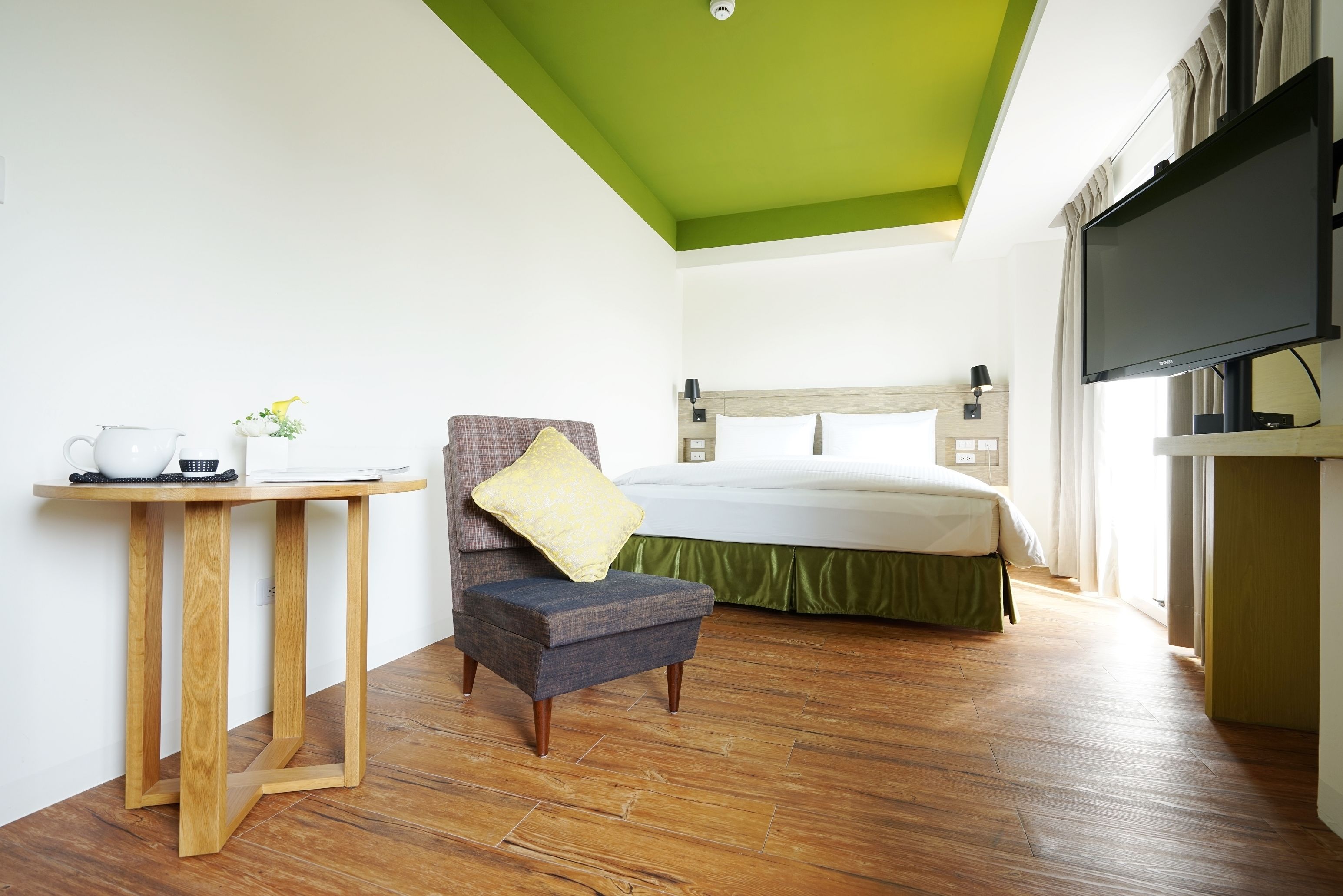 A hotel room with a bed, a television, a small table with a kettle, and a chair.