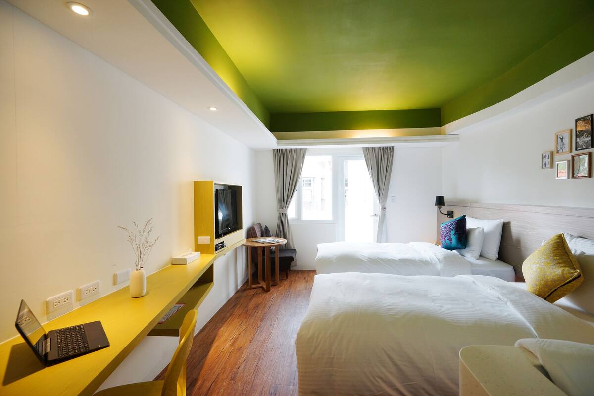 Quarto de hotel com duas camas, uma escrivaninha com um laptop, televisão e janela com cortinas.