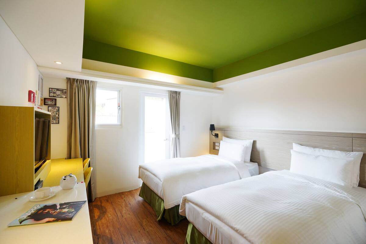 Quarto de hotel com duas camas, uma escrivaninha com cadeira, uma janela com cortinas e teto verde.