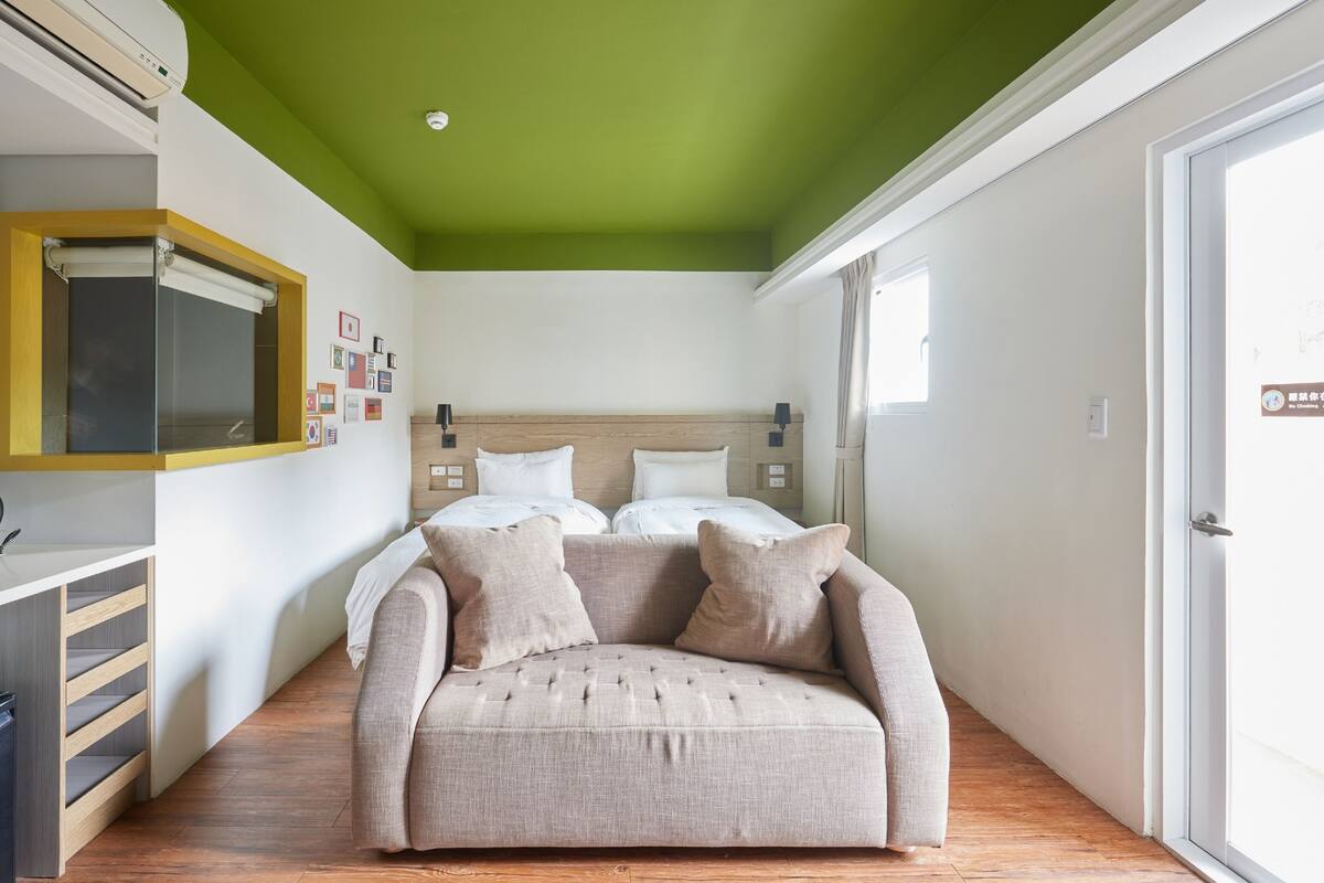 Um quarto com teto verde, uma cama com roupa de cama branca, um sofá bege e chão de madeira.