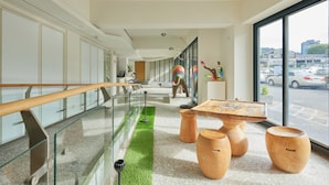 En modern hotellobby med ett glasräcke, en grön konstgräsbelagd gång, trästolar och ett stort fönster med utsikt över gatan.