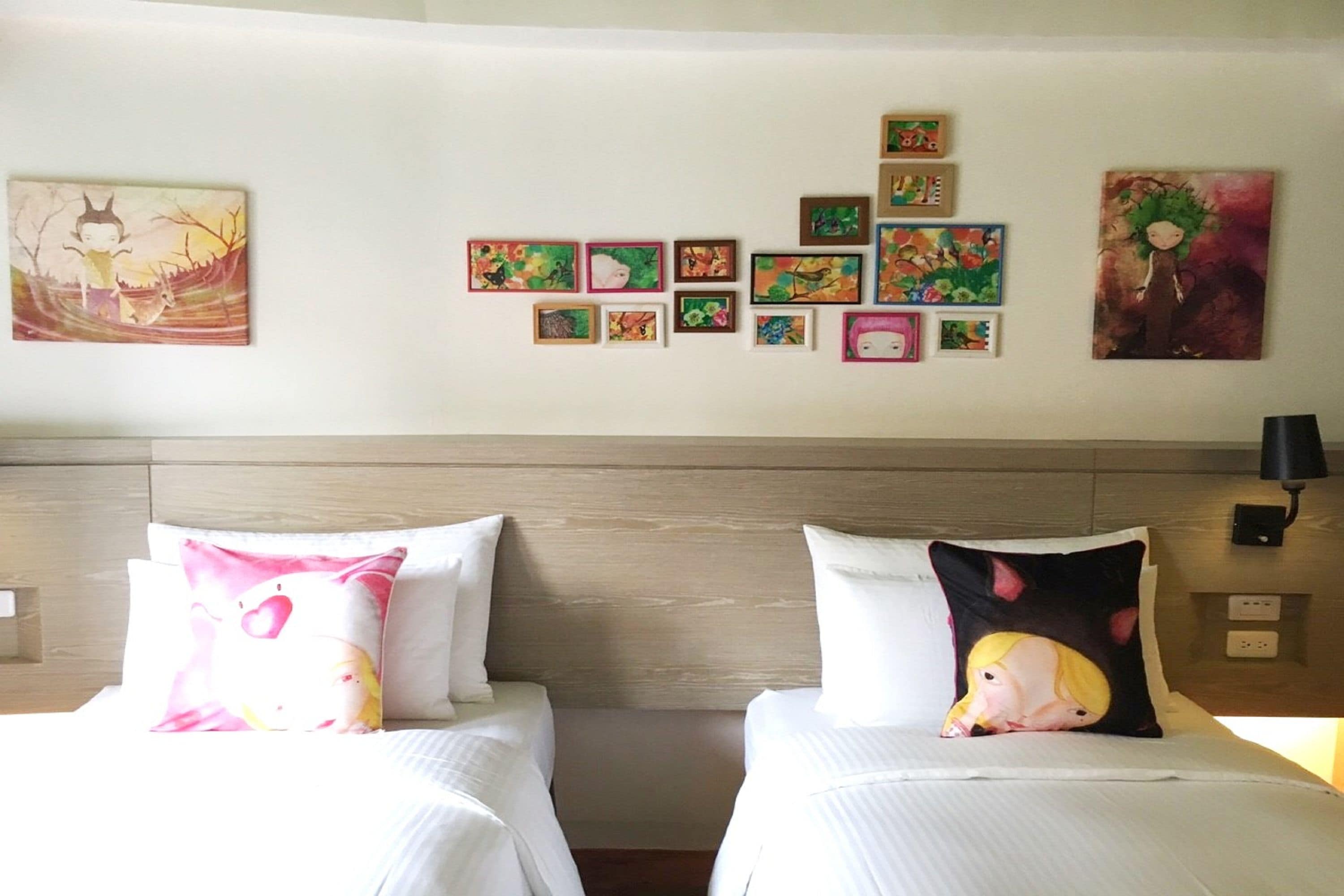 Une chambre d’hôtel avec deux lits, chacun agrémenté d’un oreiller orné d’un personnage de dessin animé. La chambre est dotée d’une tête de lit en bois, d’un mur où sont accrochées plusieurs œuvres d’art encadrées, et d’une table de chevet avec une lampe.