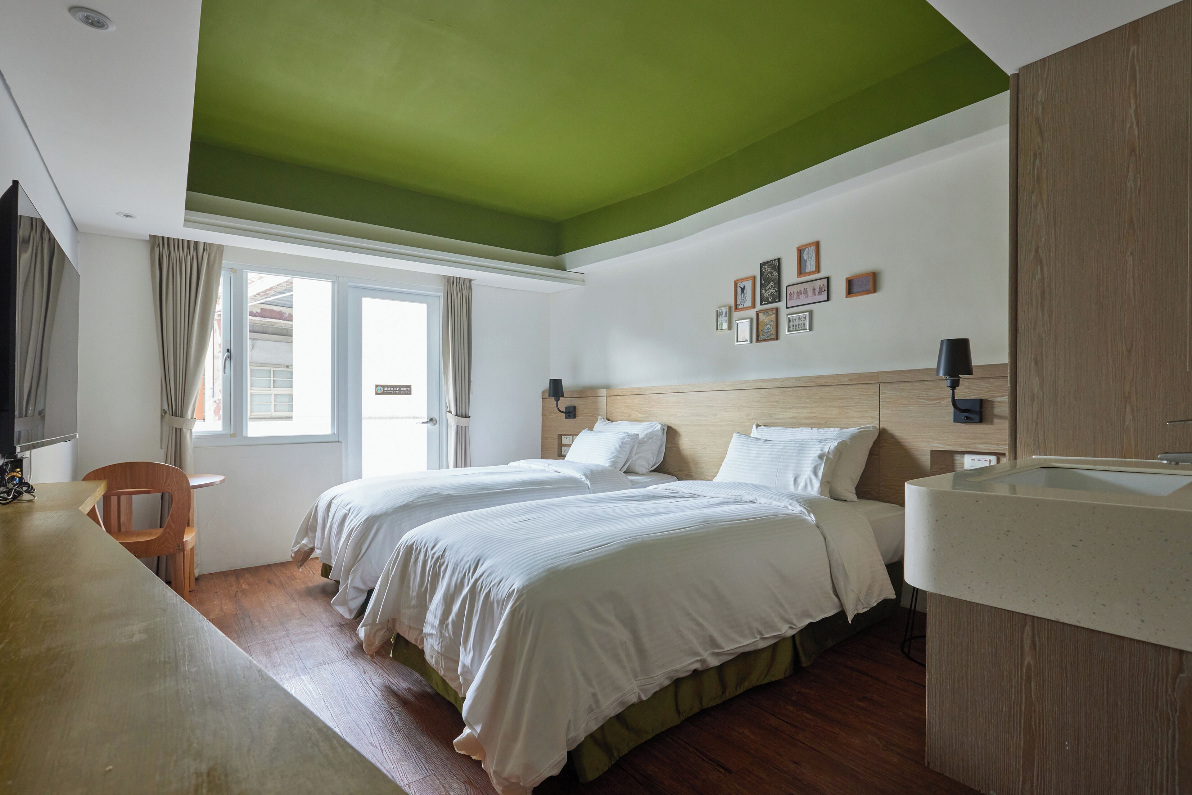 A hotel room with two beds, a wooden desk, a large window with curtains, and a green ceiling.