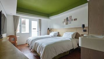 A hotel room with two beds, a wooden desk, a large window with curtains, and a green ceiling.