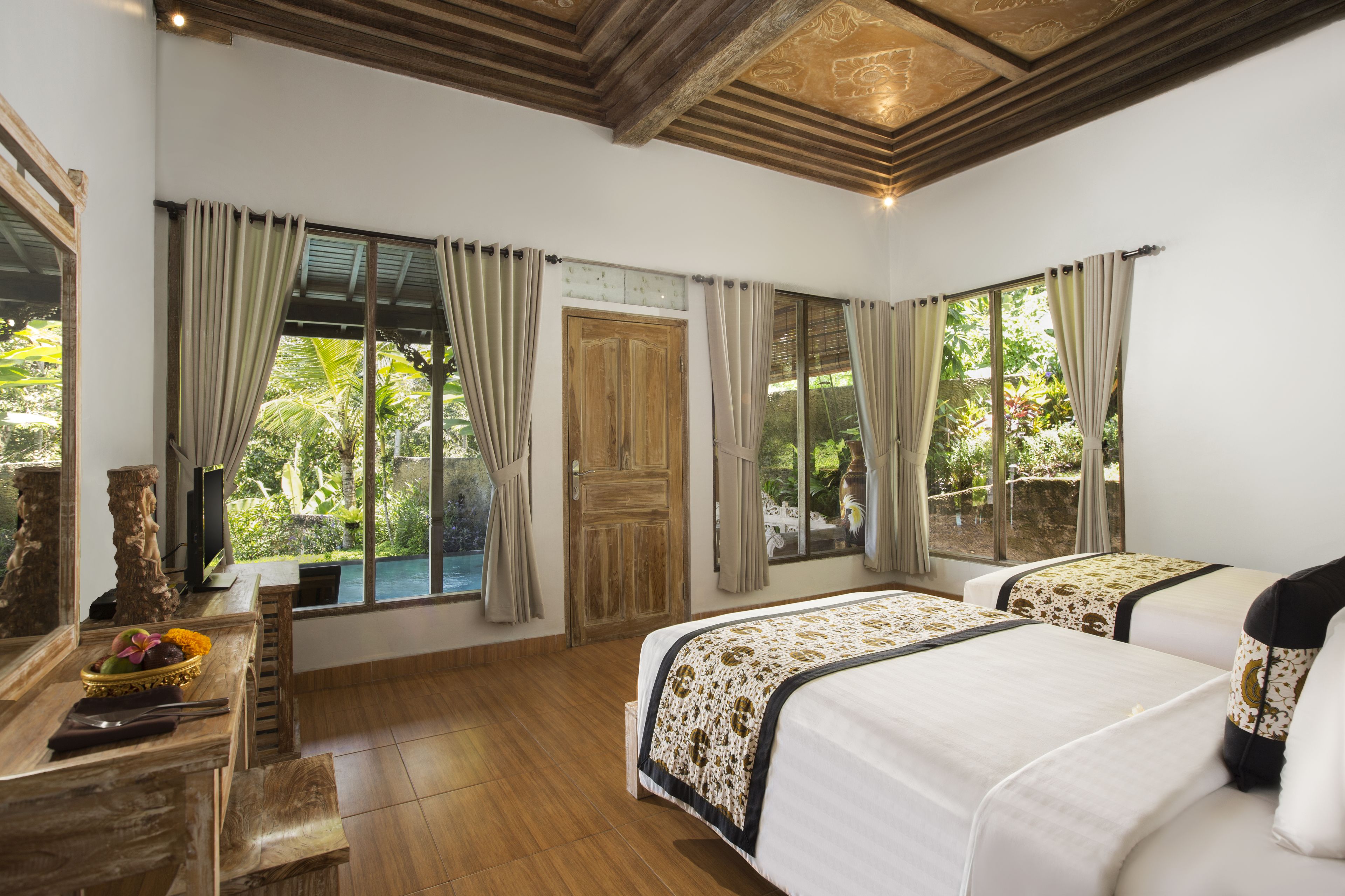 A room with two beds, wooden flooring, a wooden door, and large windows with curtains.