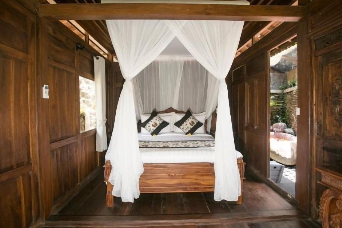 Una cama con dosel de madera, cortinas blancas, una pared de piedra y vistas a un jardín.