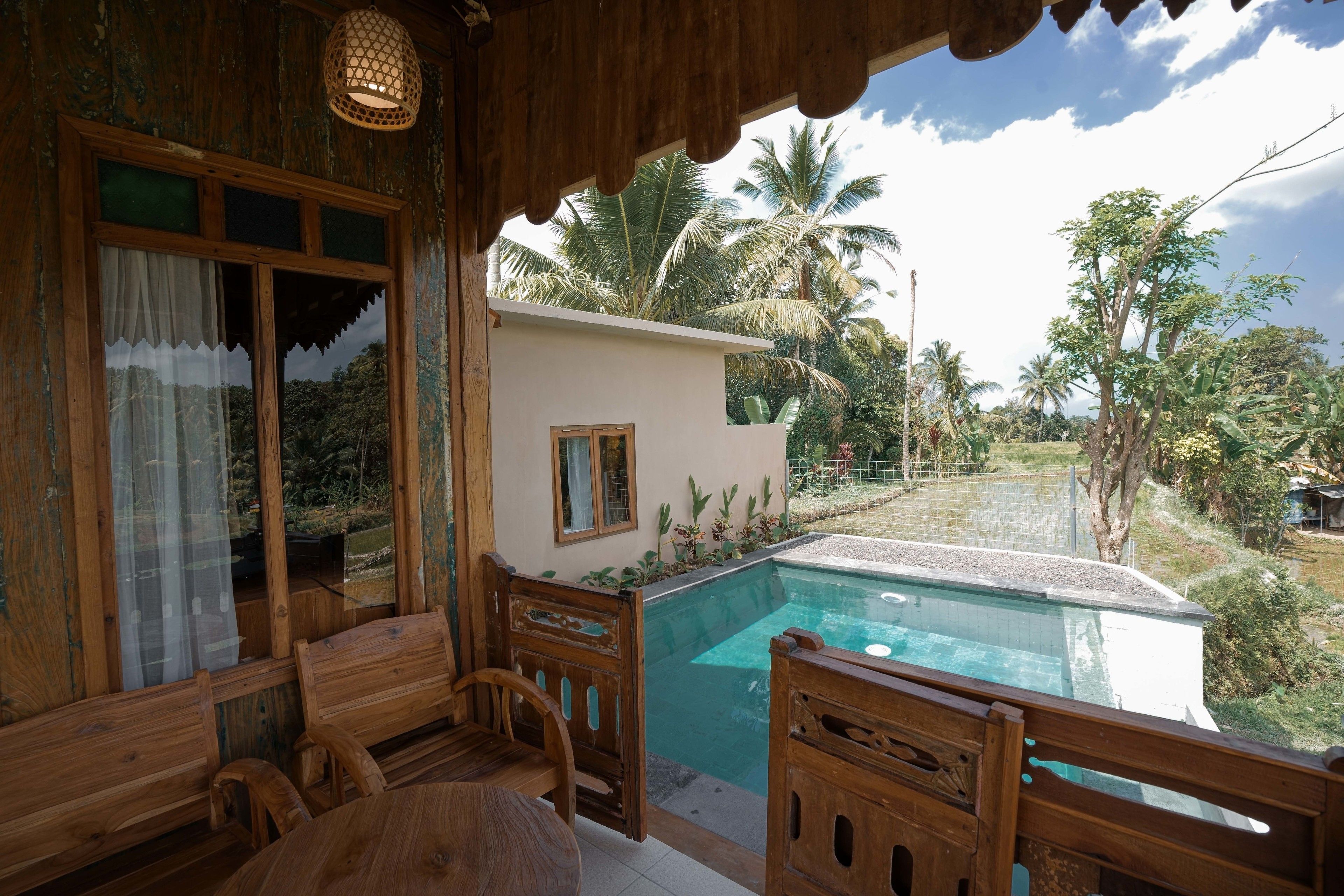 A wooden deck with a pool, wooden furniture, and a view of palm trees and greenery.