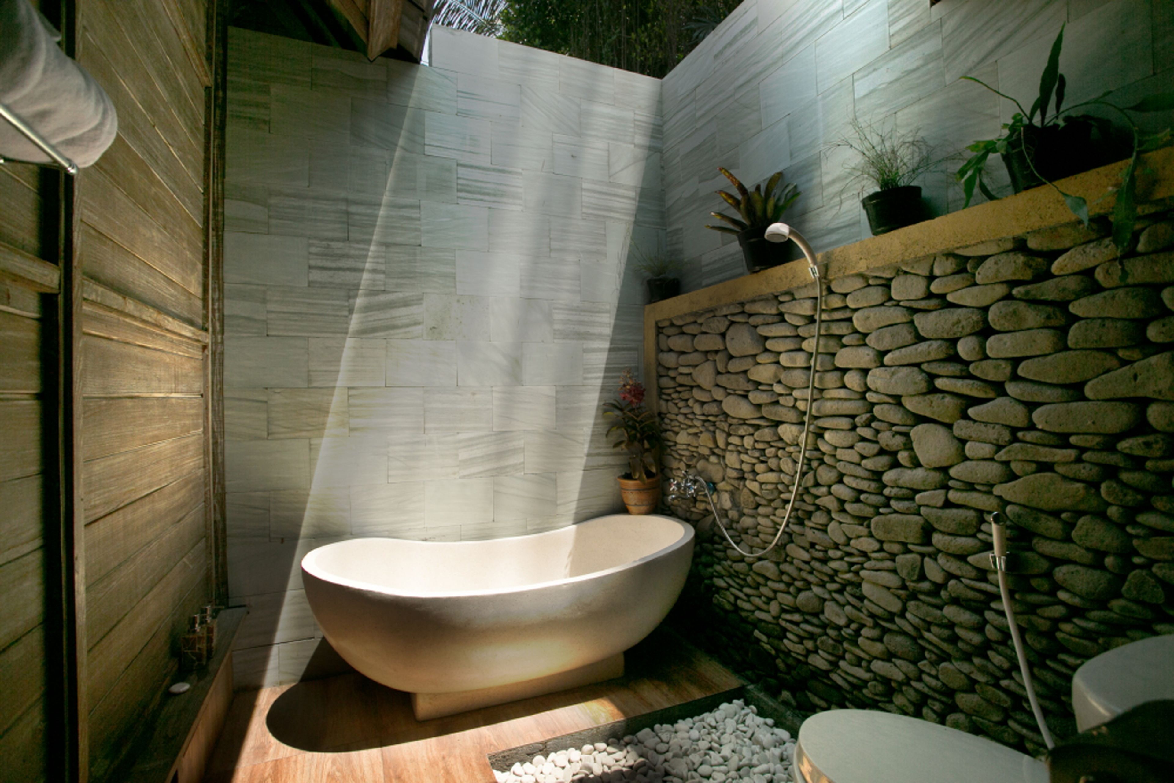 A modern bathroom with a freestanding bathtub, stone wall, and potted plants.