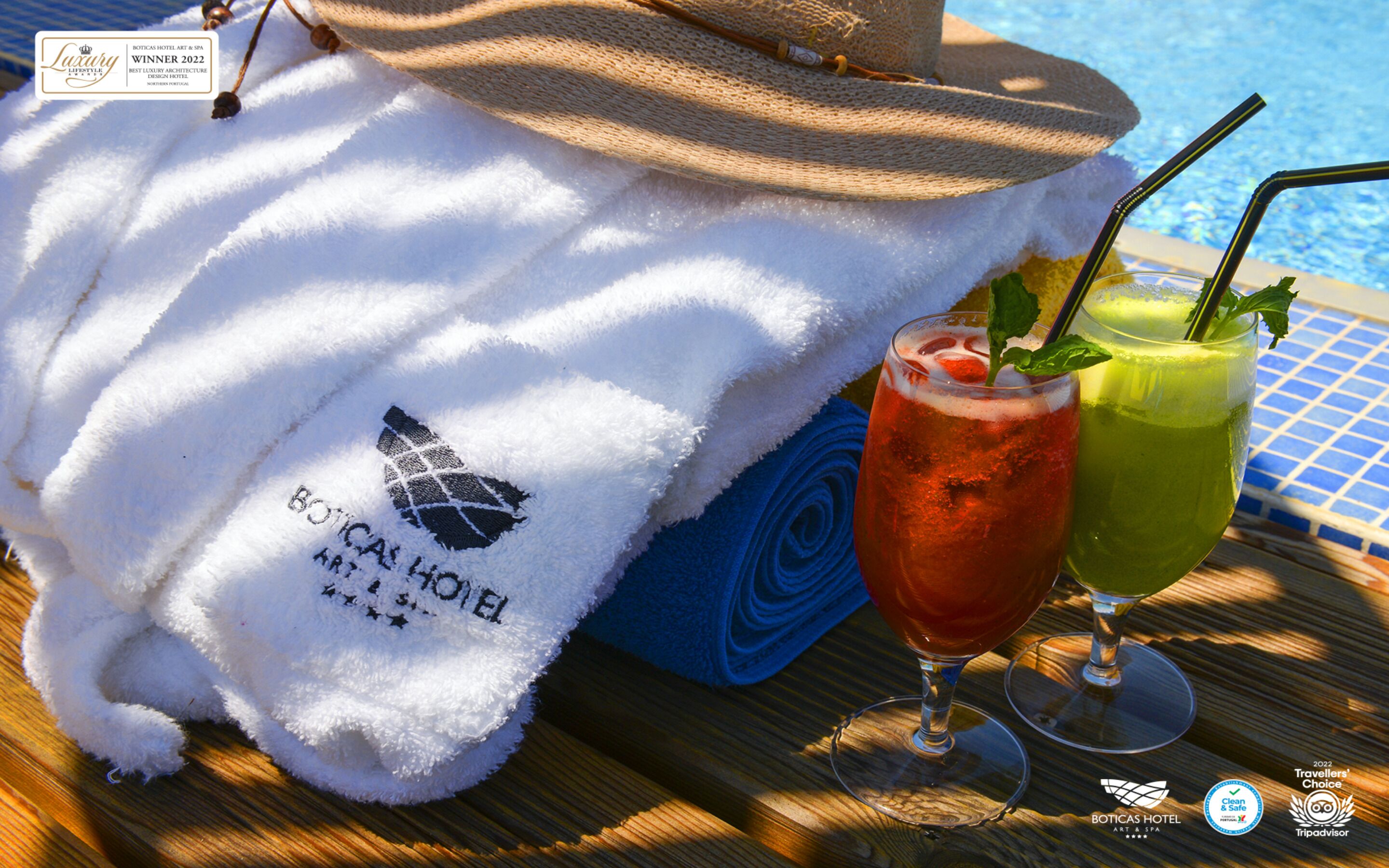 A towel with the BOTICAS HOTEL logo, a straw in a glass of red drink, and a straw in a glass of green drink on a wooden table by a pool.