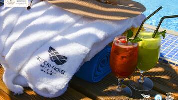 A towel with the BOTICAS HOTEL logo, a straw in a glass of red drink, and a straw in a glass of green drink on a wooden table by a pool.