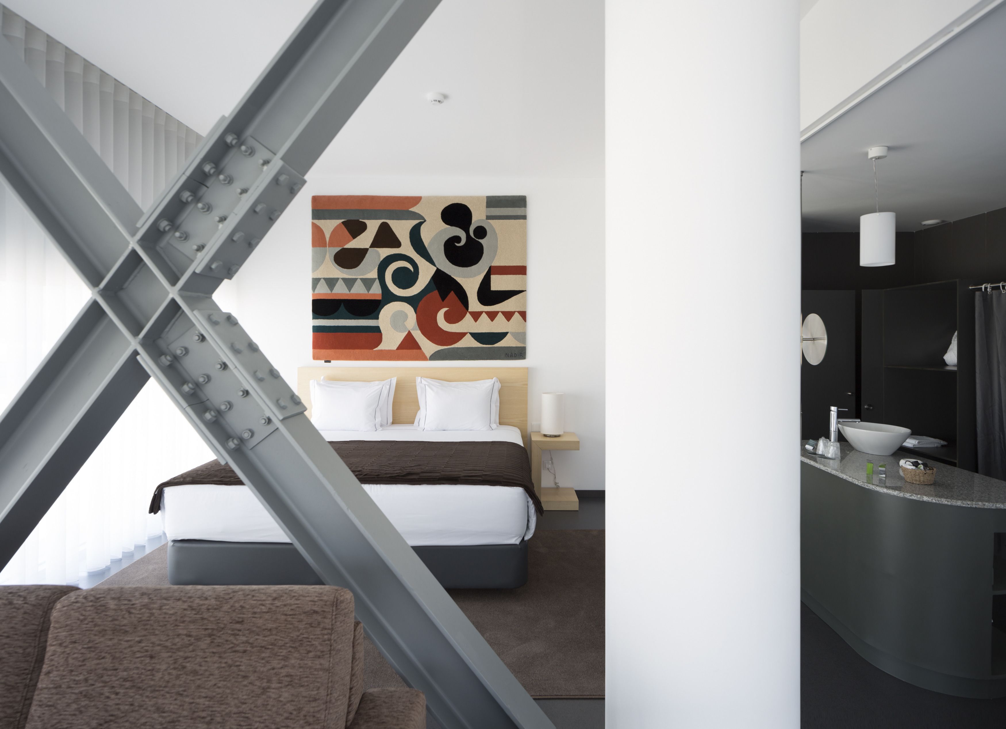 Una camera d'hotel moderna con un letto, un comodino, un quadro astratto di grandi dimensioni e un bagno con lavandino e specchio.