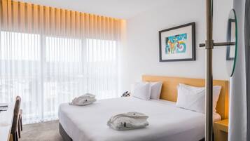 A hotel room with a large bed, a bedside table, a chair, and a window with sheer curtains.