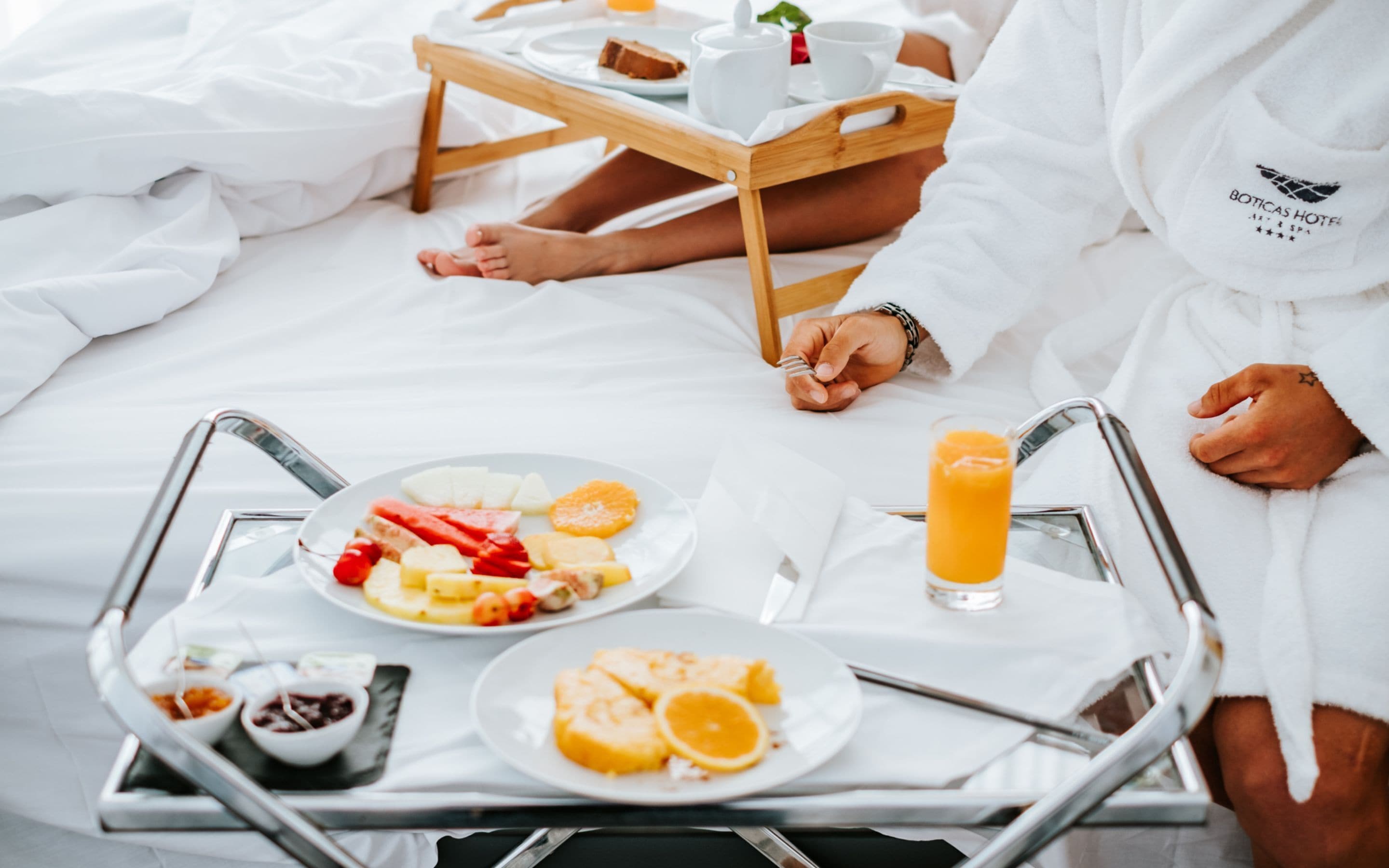 A breakfast tray with fruits, a glass of orange juice, and a cup on a bed.