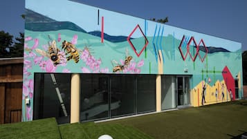 Un edificio moderno con un mural de abejas y flores, grandes ventanales de vidrio y un área verde frente a él.
