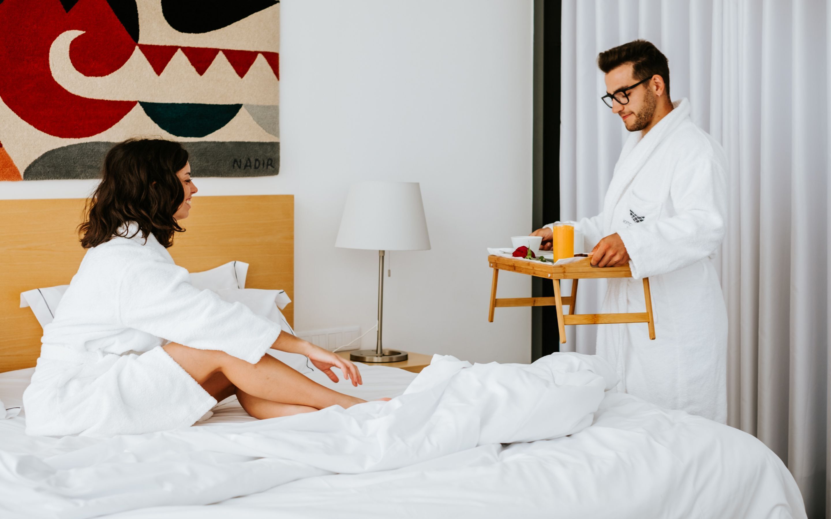 A hotel room with a bed, a bedside table with a lamp, and a wall with a colorful abstract painting.