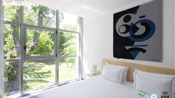 A hotel room with a large window, a bed with white linens, a modern abstract painting, and several awards displayed on the wall.