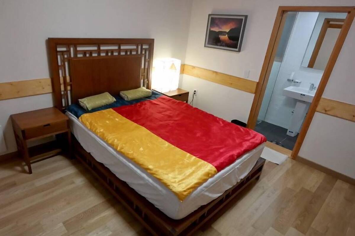 Quarto com cama de madeira, mesa de cabeceira, espelho e banheiro visível por uma porta aberta.