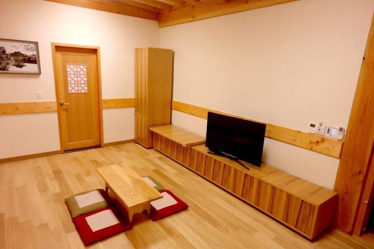 Um quarto pequeno e acolhedor com piso de madeira, uma TV sobre um móvel baixo de madeira, uma porta com painel de vidro decorativo e um quadro na parede.