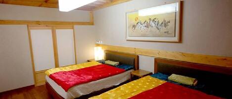 A room with two beds, wooden beams on the ceiling, a framed artwork on the wall, and a bedside table with a lamp.