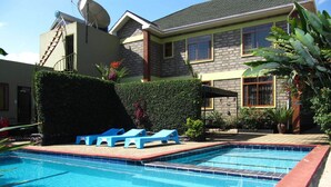 A swimming pool with blue lounge chairs, a brick border, and a house with a stone facade in the background. - Korona Villa Bed & Breakfast (Arusha)