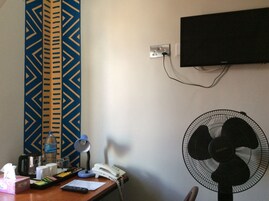 Habitación de hotel con escritorio, teléfono, ventilador, televisor montado en la pared y un tapiz decorativo en la pared.