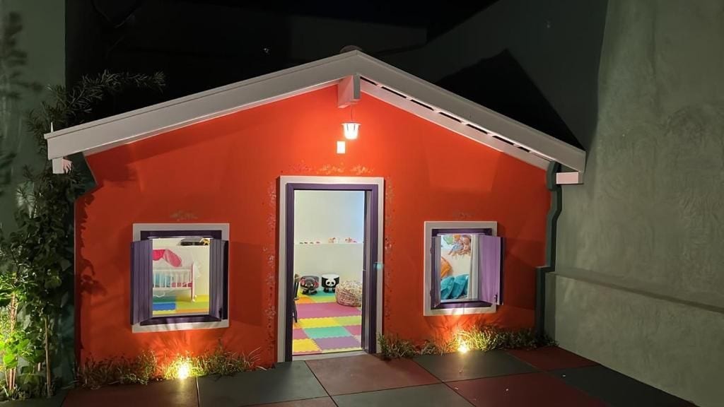 Un edificio pequeño, con mucha luz, de fachada roja, una puerta y dos cuadros en cada lado.
