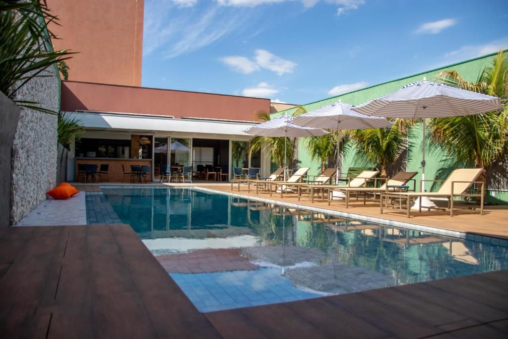 A swimming pool with lounge chairs and umbrellas, surrounded by palm trees and a building with a sloped roof.