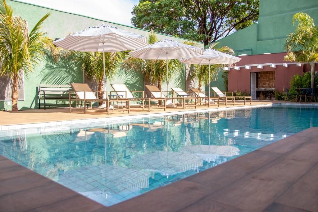 Piscina con sillones de descanso y sombrillas, rodeada de vegetación y un edificio de fachada roja.