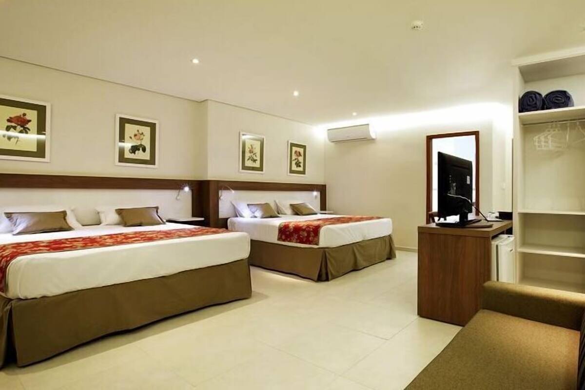 A hotel room with two beds, a desk with a computer, a wardrobe, and framed pictures on the wall.
