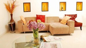 Un salon avec un canapé beige, une table basse en verre sur laquelle sont posés des magazines, et un vase rempli de fleurs. Des tableaux encadrés ornent le mur.