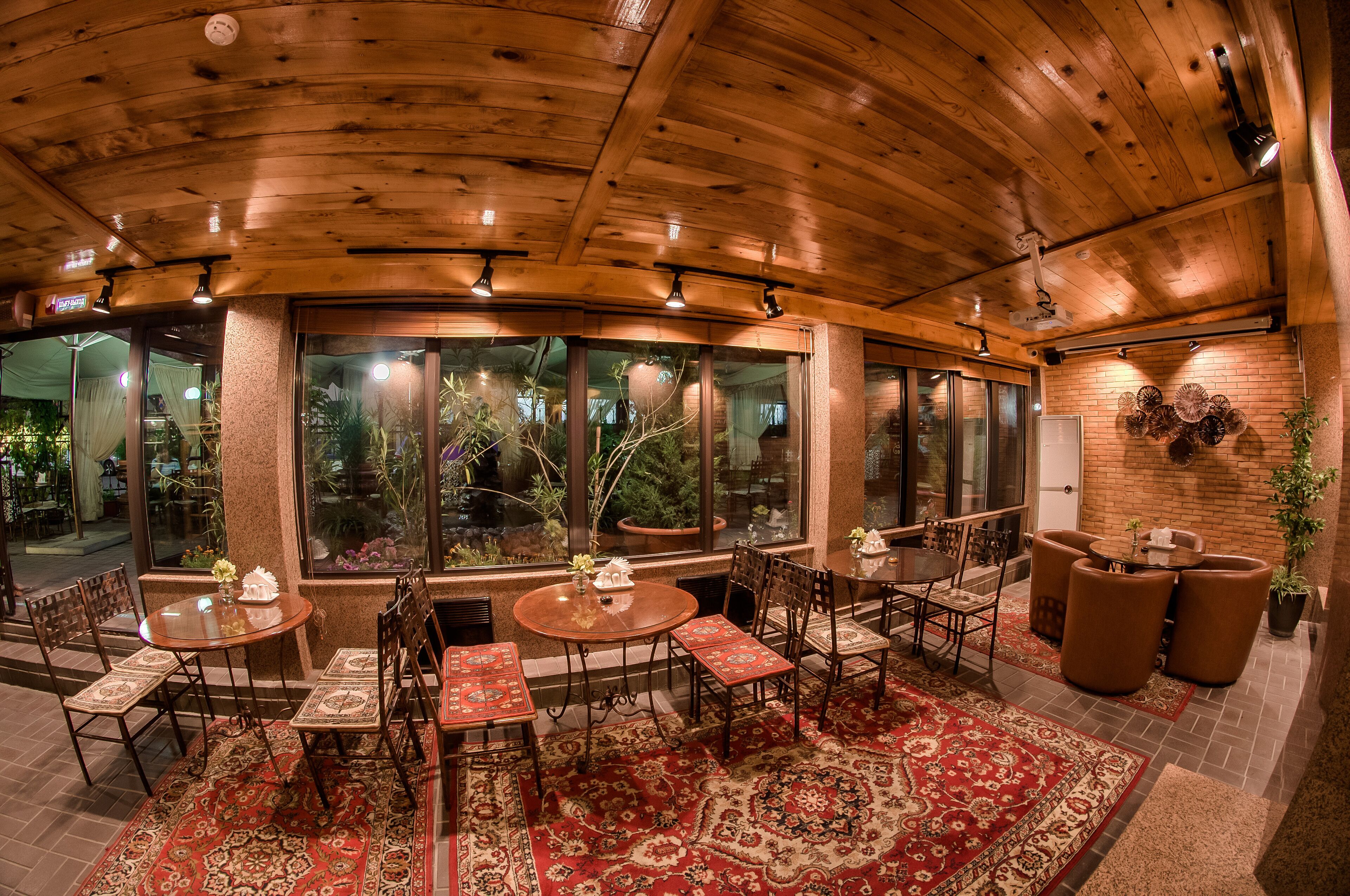 A cozy restaurant with wooden ceilings, large windows, and a variety of seating arrangements including round tables and cushioned chairs.