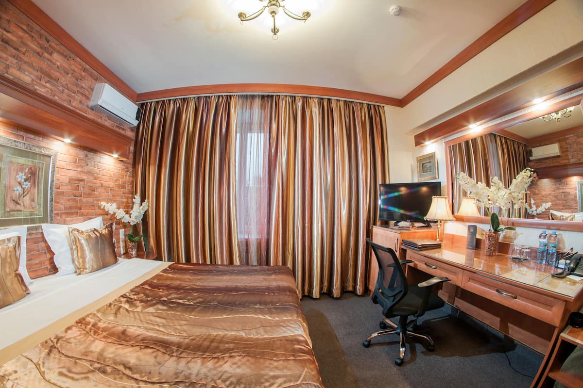 A hotel room with a bed, desk, and chair, a large window with curtains, and a brick wall.
