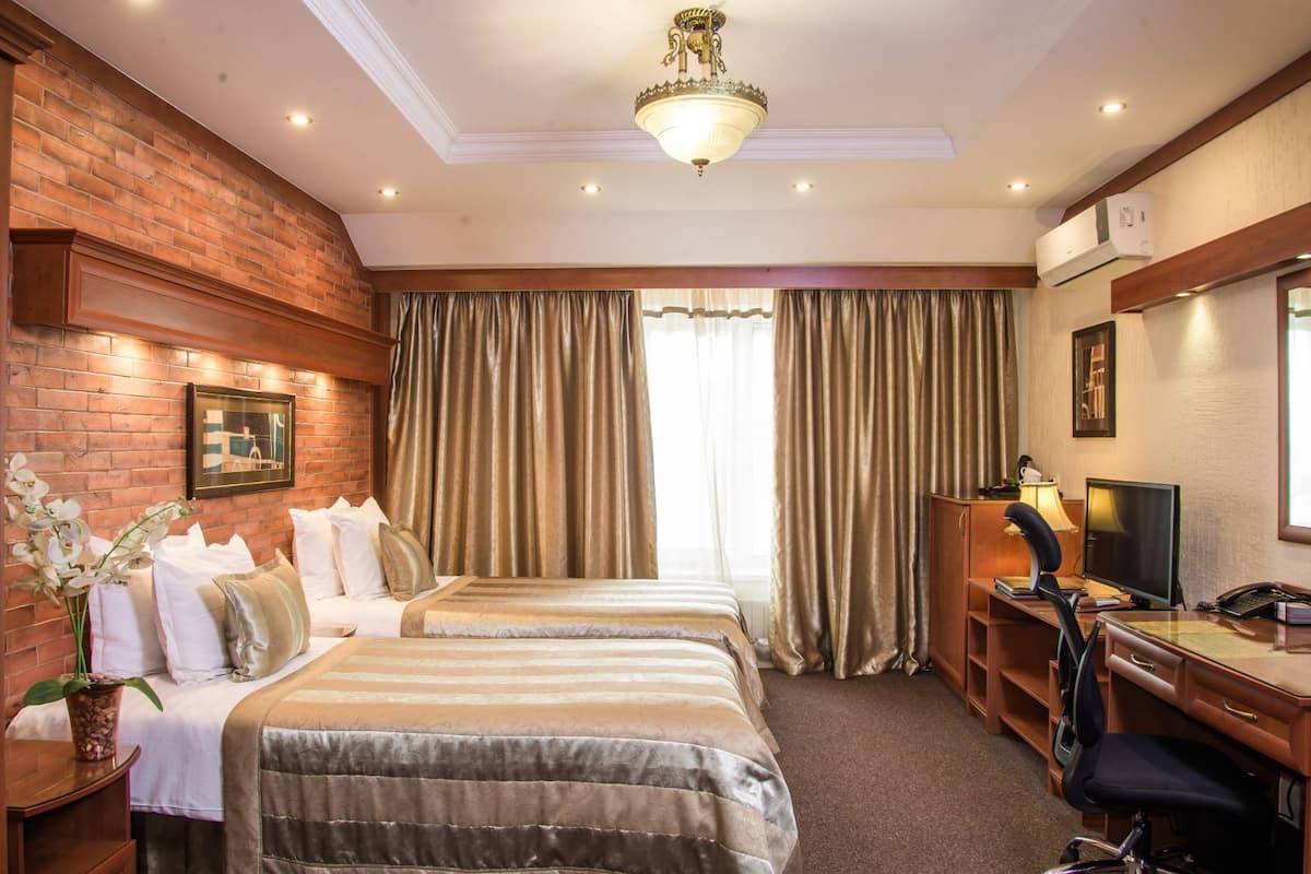 A hotel room with two beds, a desk with a computer, a chair, a desk lamp, and a large window with curtains.