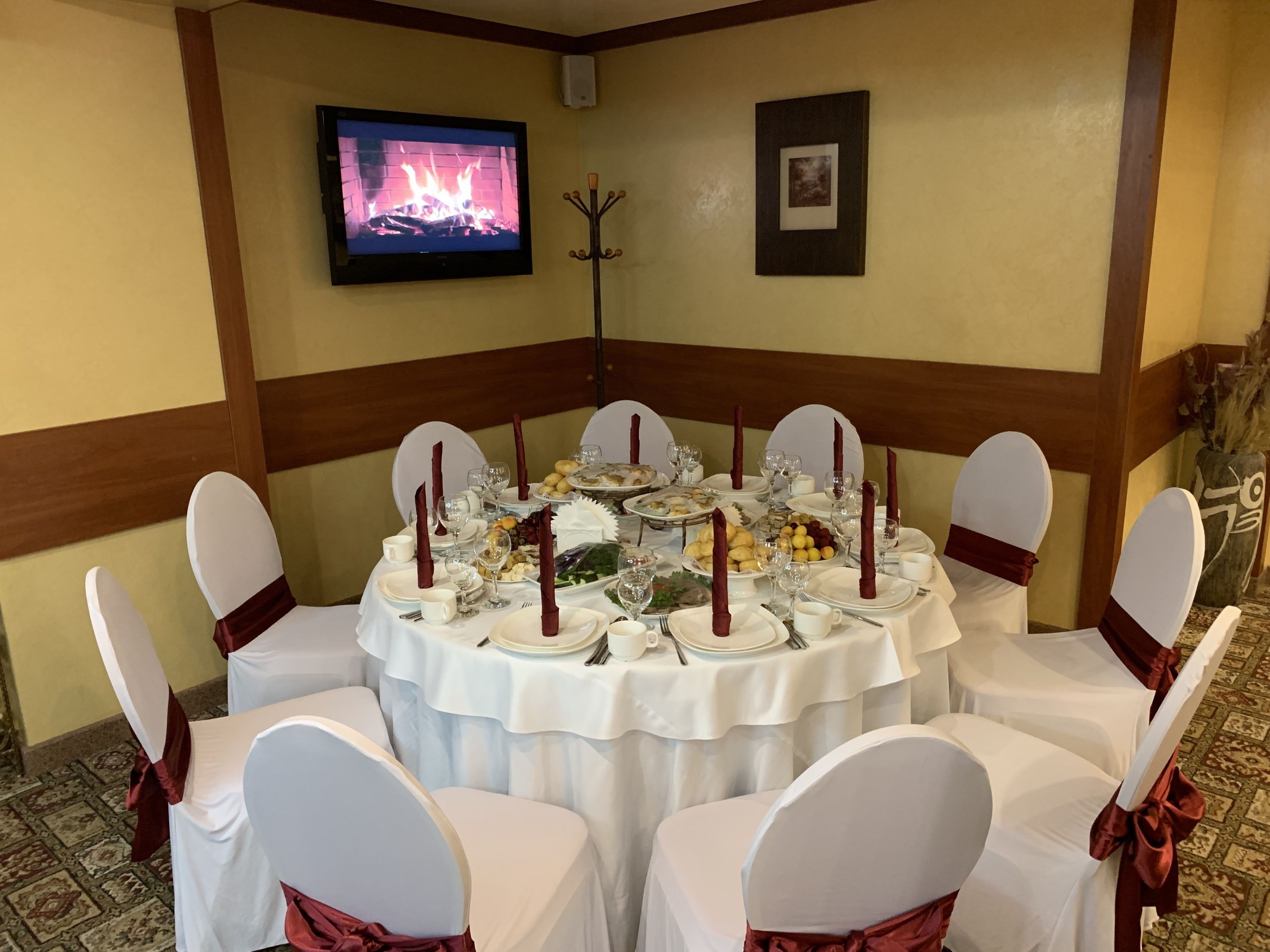 Una mesa redonda dispuesta para un evento formal, con sillas blancas y cintas rojas, un televisor mostrando una chimenea y cuadros en la pared.