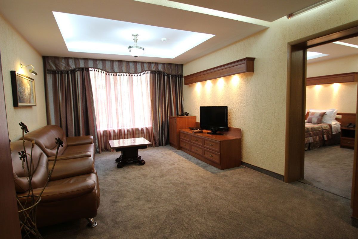 A hotel room with a sofa, a TV on a wooden cabinet, a ceiling light, and a view of another room.