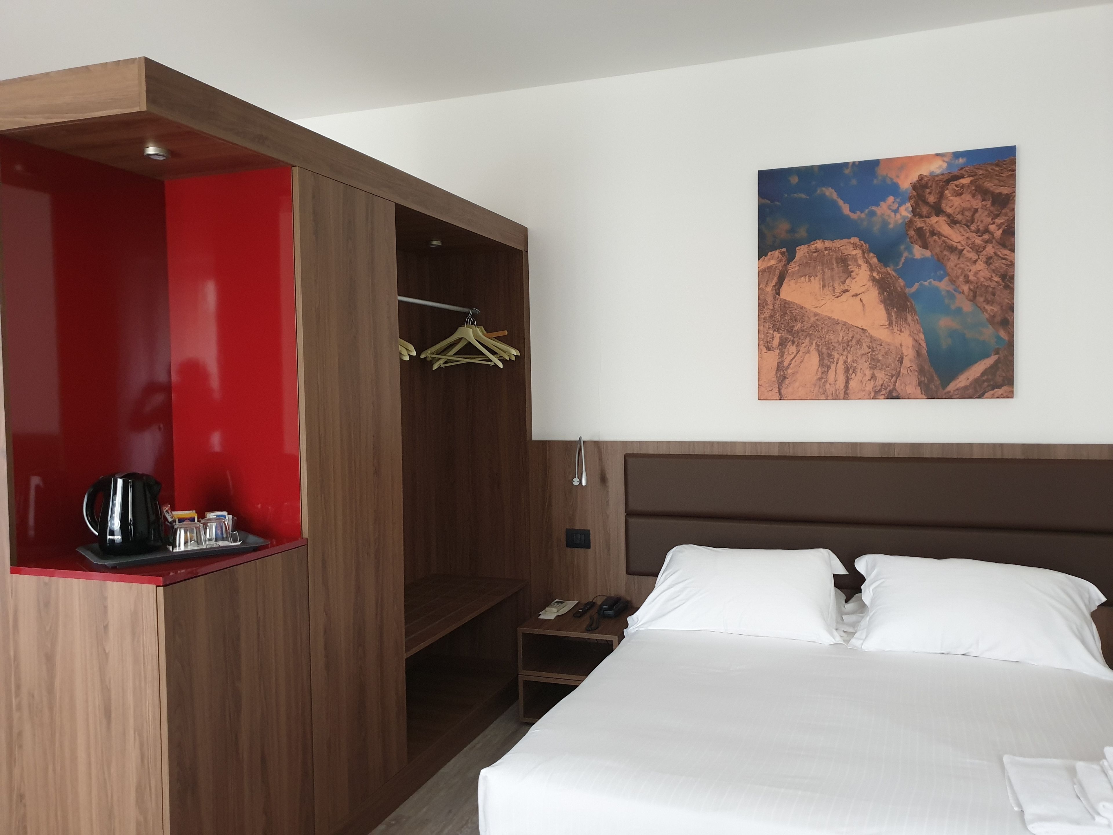 A hotel room with a bed, a red and brown wardrobe, a painting of a mountain, and a nightstand with a phone and a lamp.