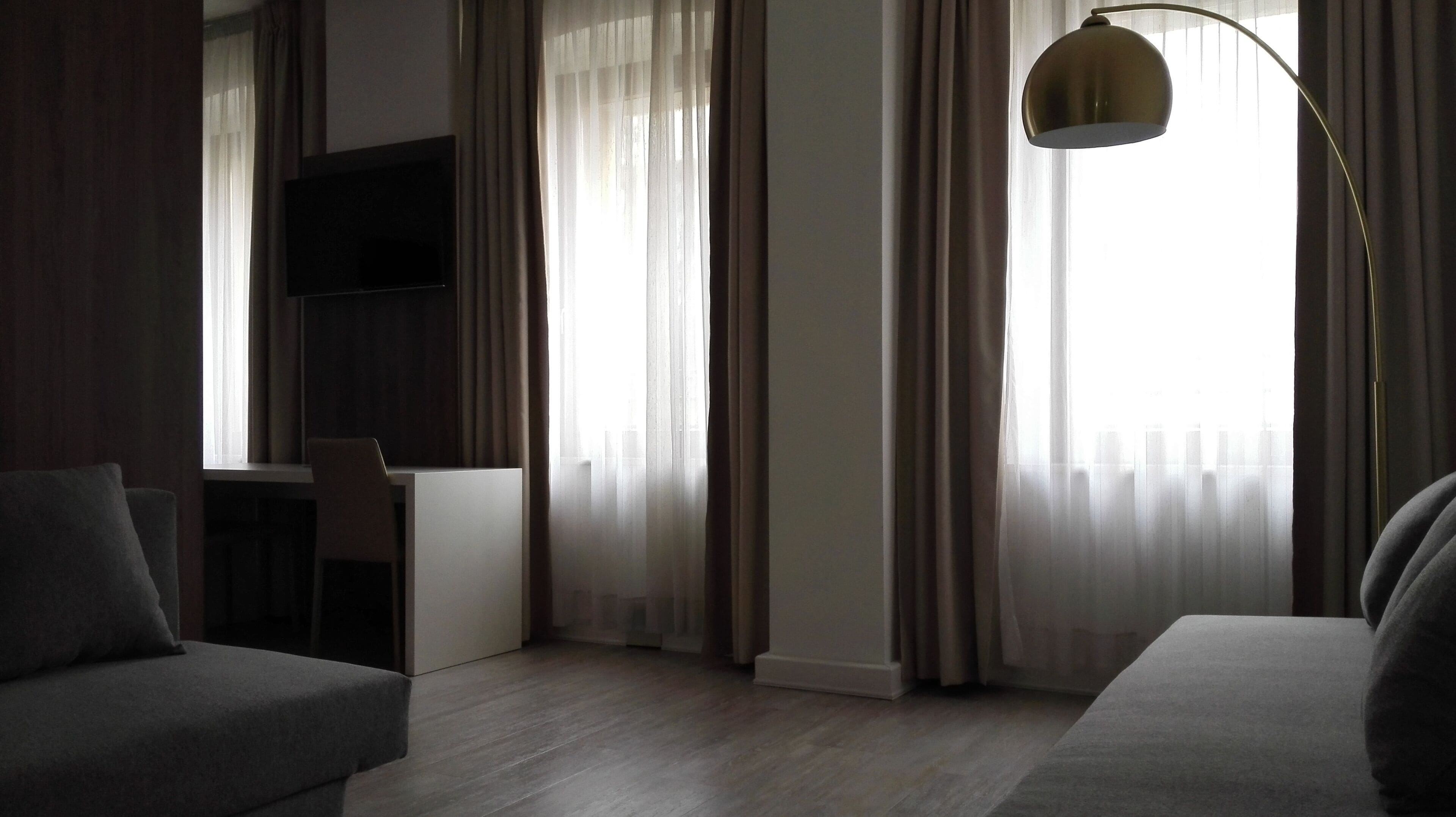 A modern hotel room with a flat-screen TV mounted on the wall, a desk, and two grey sofas.
