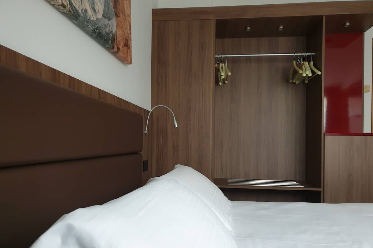 A bedroom with a wooden headboard, a white bed, and a wooden wardrobe with a mirror and hanging clothes.