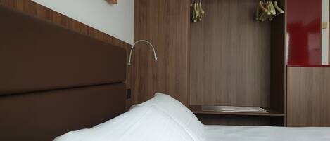 A bedroom with a wooden headboard, a white bed, and a wooden wardrobe with a mirror and hanging clothes.